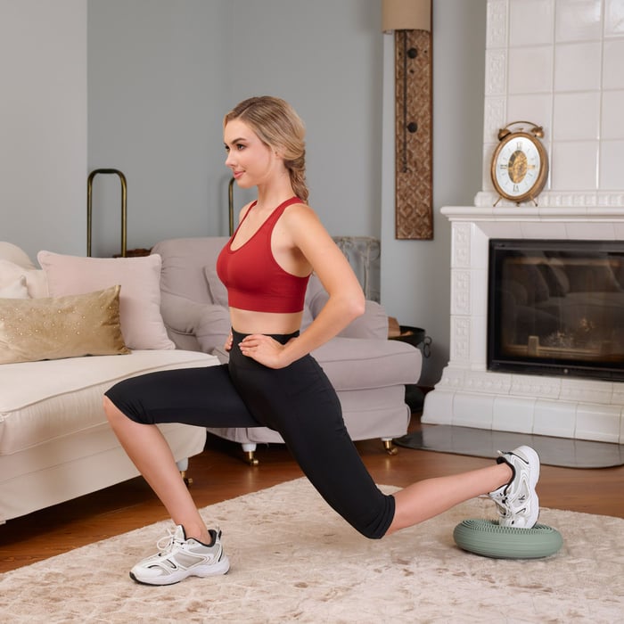 Frau macht einen Ausfallschritt und trainiert das Gleichgewicht auf einem grünen Balancekissen von Hop-Sport im Wohnzimmer.