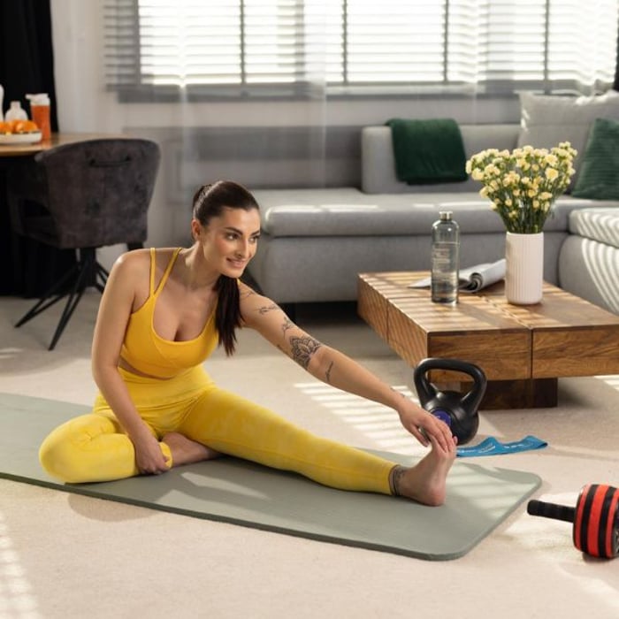 Eine Frau in gelber Sportkleidung dehnt sich auf einer grauen Yogamatte im Wohnzimmer. Neben ihr liegen eine Hop-Sport Kettlebell, ein Widerstandsband und eine Hantelstange.