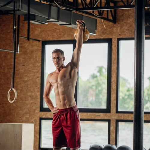 Ein Mann in einer Sporthalle führt die Übung „Kettlebell Military Press“ aus.