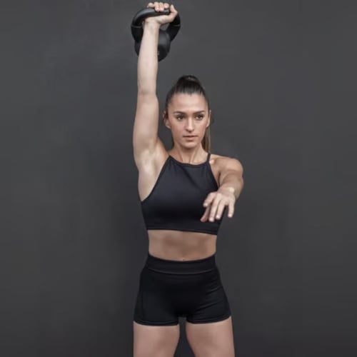 Junge Frau in schwarzem Sportoutfit hält eine Kettlebell über den Kopf beim Training, macht Kettlebell Snatch