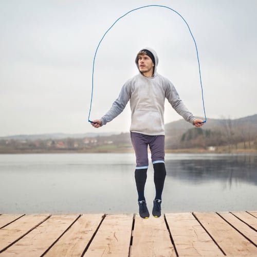 Mann trainiert mit einem Springseil auf einem Holzsteg vor einem ruhigen See. Er trägt Sportkleidung und springt in die Luft.