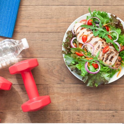 Ein Teller mit gegrilltem Hähnchen und frischem Salat, daneben eine Wasserflasche, eine rote Kurzhantel und eine blaue Yogamatte auf einem Holztisch.