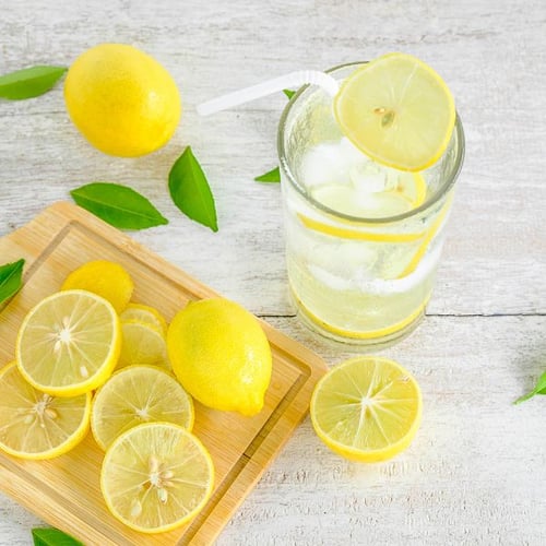 Ein Glas Zitronenwasser mit Eiswürfeln und einer Zitronenscheibe auf dem Rand steht neben einer Holzplatte mit mehreren geschnittenen Zitronenscheiben. Ganze Zitronen und grüne Blätter liegen auf dem Tisch.