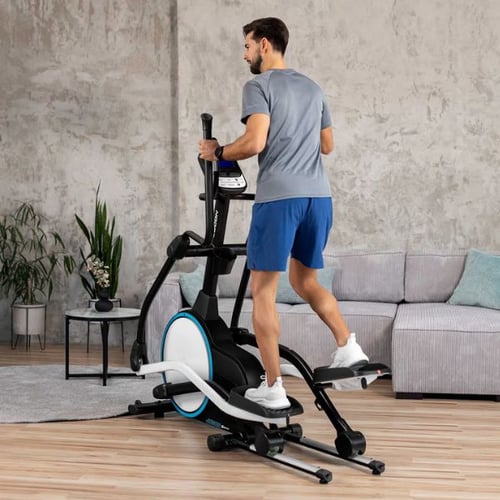 Ein Mann trainiert auf einem Hop-Sport Crosstrainer in einem modernen Wohnzimmer. Der Crosstrainer hat ein großes Schwungrad mit blauer Verzierung und stabile Haltegriffe. 