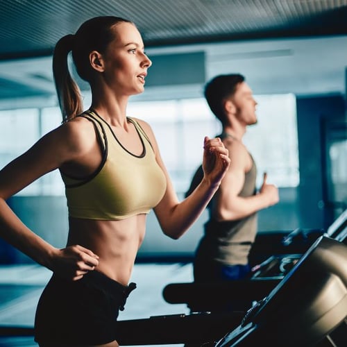 Zwei Personen laufen auf Laufbändern in einem Fitnessstudio. Im Vordergrund ist eine Frau mit einem gelben Sport-BH zu sehen, die konzentriert läuft. Ein Mann läuft im Hintergrund.