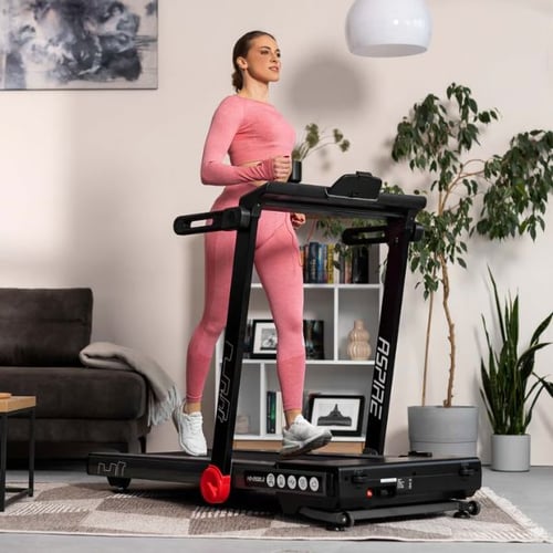 Frau auf dem Hop-Sport Aspire Laufband in einem Wohnzimmer. Sie trägt ein rosa Trainingsoutfit und läuft entspannt. Im Hintergrund sind eine Couch, ein Bücherregal und eine Pflanze zu sehen.