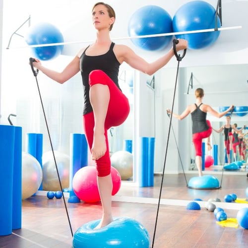 Frau trainiert mit einem blauen Balance-Trainer und Fitnessbändern in einem hellen Trainingsraum.