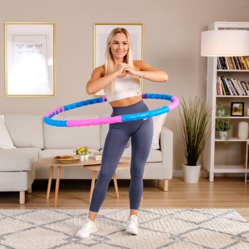 Eine Frau in einem gemütlichen Wohnzimmer nutzt einen blau-pinken Hop-Sport Fitness-Hula-Hoop-Reifen für ihr Training. Sie trägt graue Leggings und ein weißes Sporttop, während sie den Reifen in Bewegung hält. Im Hintergrund sind ein Sofa, ein Couchtisch und Bücherregale zu erkennen.