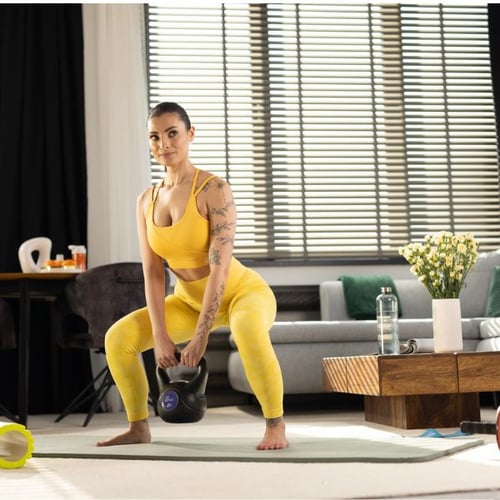 Frau im gelben Sportoutfit macht ein Training mit einer schwarzen Hop-Sport Kettlebell in einem Wohnzimmer. Sie hält die Kettlebell fest und ist in einer Kniebeuge-Position.