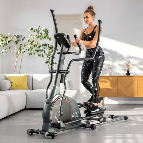 Frau trainiert auf einem Hop-Sport Crosstrainer Prim, in einem modernen Wohnzimmer mit hellem Sofa und Pflanzen im Hintergrund.