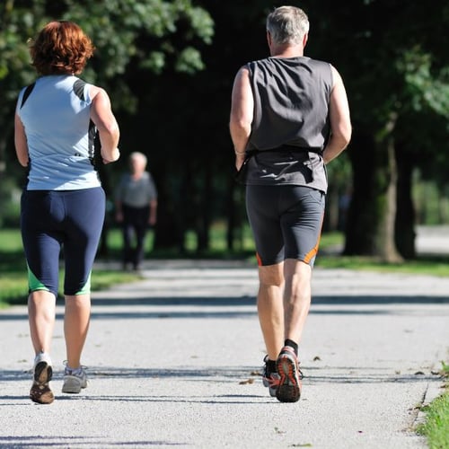 Zwei Personen, ein Mann und eine Frau, slow joggen auf einem sonnigen Parkweg. Sie tragen sportliche Kleidung und sind von hinten zu sehen.