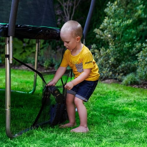 Ein kleiner Junge legt seine Sandalen in die Schuhaufbewahrungstasche eines Hop-Sport Trampolins im Garten