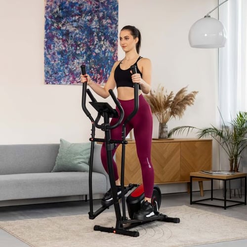 Frau auf einem Hop-Sport Crosstrainer in einem modernen Wohnzimmer, trainiert in sportlicher Kleidung.