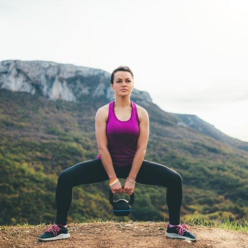 Frau macht ein Kettlebell-Sumo-Squat-Training in der Natur mit Bergen im Hintergrund.