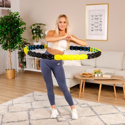 Eine Frau in einem modernen Wohnzimmer trainiert mit einem gelb-schwarzen Hop-Sport Fitness-Hula-Hoop-Reifen. Sie trägt ein weißes Sporttop und graue Leggings, während sie den Reifen um ihre Hüfte schwingt. Im Hintergrund sind ein Sofa, ein Couchtisch und Pflanzen zu sehen.