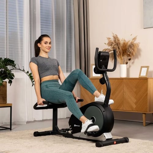 Frau auf einem Hop-Sport Liegeergometer in einem gemütlichen Raum mit Deko und Holzkommode im Hintergrund.