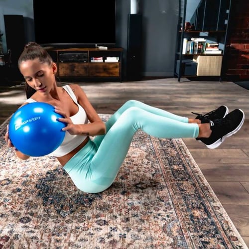 Frau in hellblauer Sportkleidung hält einen blauen Hop-Sport Pilatesball und macht eine Rumpfrotation auf einem Teppich.