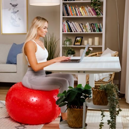 Frau sitzt auf rotem Gymnastikball mit Noppen am Schreibtisch und arbeitet am Laptop