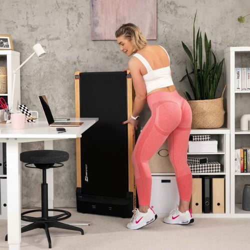 Eine Frau verstaut das Hop-Sport Laufband Primo senkrecht gegen die Wand in einem Büro-Wohnzimmer. Das Laufband ist platzsparend zusammengeklappt.