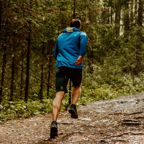 Mann in blauer Jacke joggt auf einem Waldweg bei nassem Wetter.