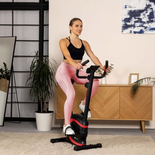 Frau im schwarzen Sporttop und pinker Leggings trainiert auf einem Hop-Sport Heimtrainer Vox in einem modernen Wohnzimmer. Sie sitzt aufrecht und lächelt während des Trainings.