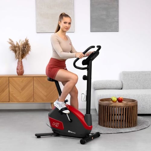 Eine junge Frau trainiert auf einem roten Hop-Sport Heimtrainer in einem modernen Wohnzimmer. Sie trägt ein beiges Oberteil und rote Shorts. Im Hintergrund steht ein Sideboard mit einer Vase und Dekoration.