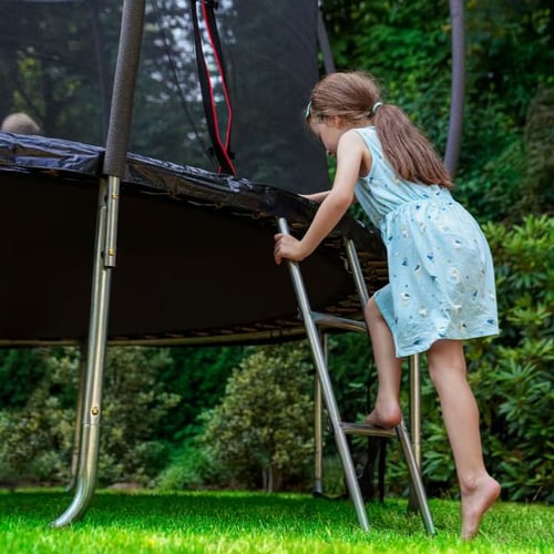 Ein Mädchen in einem Sommerkleid klettert über eine Leiter auf ein Hop-Sport Trampolin.