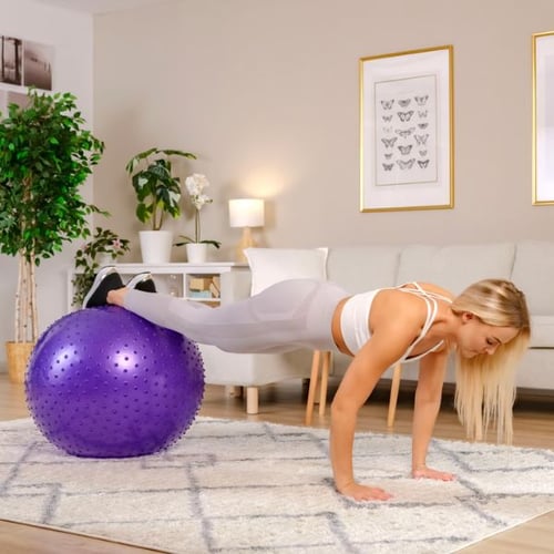 Frau führt eine Fitnessübung in einem Wohnzimmer durch. Ihre Füße ruhen auf einem lila Gymnastikball von Hop-Sport mit Massage-Noppen, während sie in der Plank-Position ist.