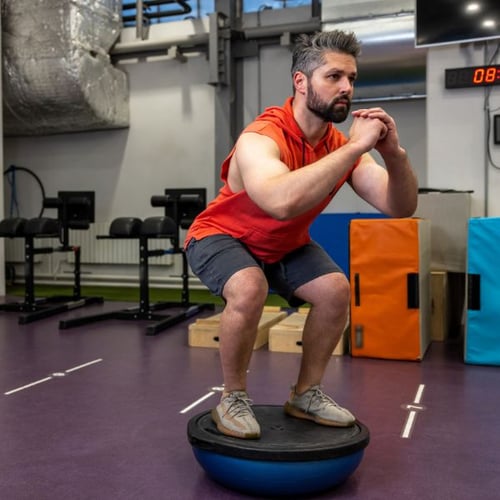 Mann führt Kniebeugen auf einem blauen Balance-Trainer in einem Fitnessstudio aus.