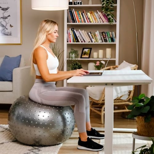 Eine Frau sitzt auf einem silbernen Hop-Sport Gymnastikball mit Noppen und arbeitet an einem Laptop. Der Ball wird als ergonomische Sitzgelegenheit in einem modernen, gemütlichen Raum verwendet.