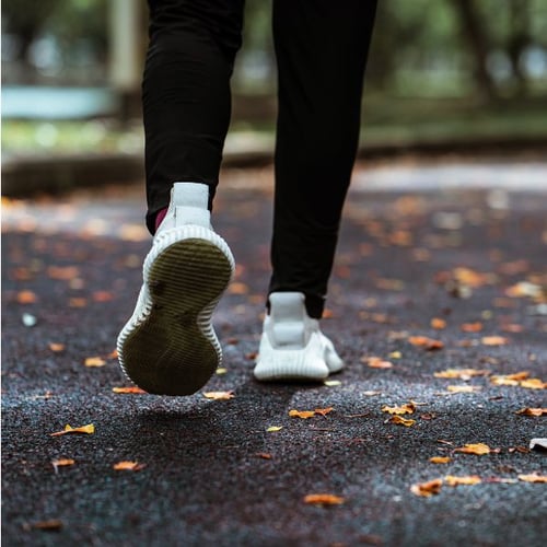 Nahaufnahme der Füße einer Person, die auf einem herbstlichen Parkweg läuft. Die Person trägt weiße Sportschuhe und schwarze Sportkleidung.