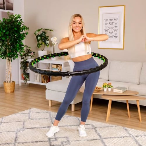 Eine Frau trainiert in einem hellen Wohnzimmer mit einem schwarz-grünen Hop-Sport Fitness-Hula-Hoop-Reifen. Sie trägt ein weißes Sporttop und enge graue Leggings. Hinter ihr sind ein Sofa, ein Couchtisch und Pflanzen zu sehen, die das Wohnzimmer dekorieren.