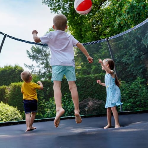 Drei Kinder spielen mit einem Ball auf einem Hop-Sport-Trampolin mit Netz in einem grünen Garten.