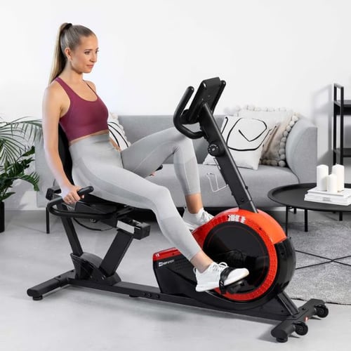 Frau trainiert auf einem Hop-Sport Liegeergometer mit rotem Schwungrad in modernem Wohnzimmer