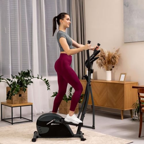 Frau trainiert in einem modernen Wohnzimmer auf einem Hop-Sport Crosstrainer Focus. Sie trägt ein graues Oberteil und weinrote Leggings und hält die Griffe des Geräts fest.