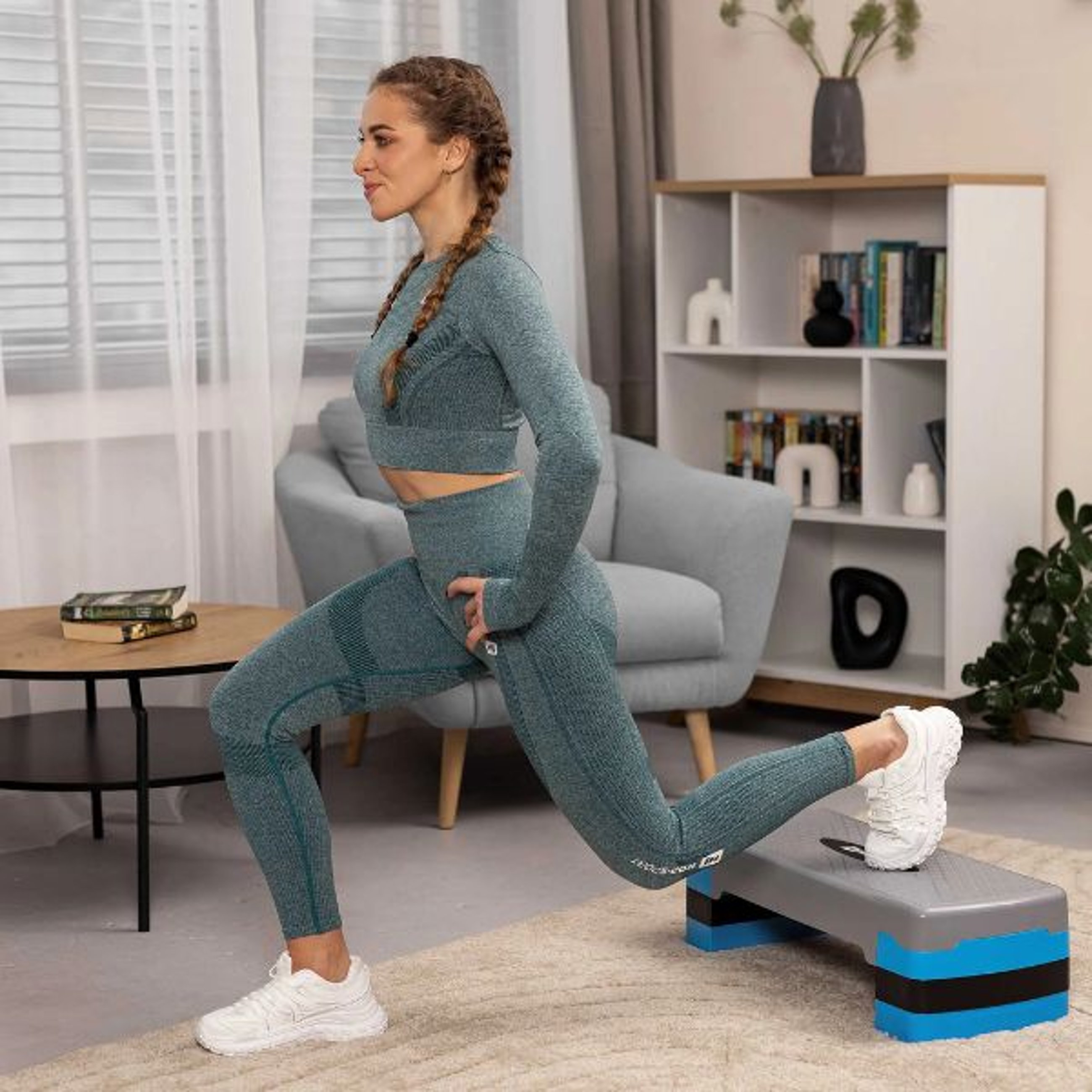 Eine Frau mit geflochtenen Haaren macht einen Ausfallschritt auf einem Hop-Sport Aerobic Stepper in Blau und Schwarz in einem Wohnzimmer.