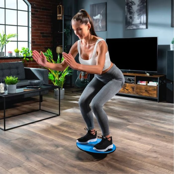 Eine Frau in Sportkleidung macht Kniebeugen auf einem blauen Hop-Sport Balance-Board. Sie hält die Arme vor sich gestreckt und balanciert auf dem Board. Im Hintergrund ist ein modern eingerichtetes Wohnzimmer mit Pflanzen und Möbeln zu sehen.