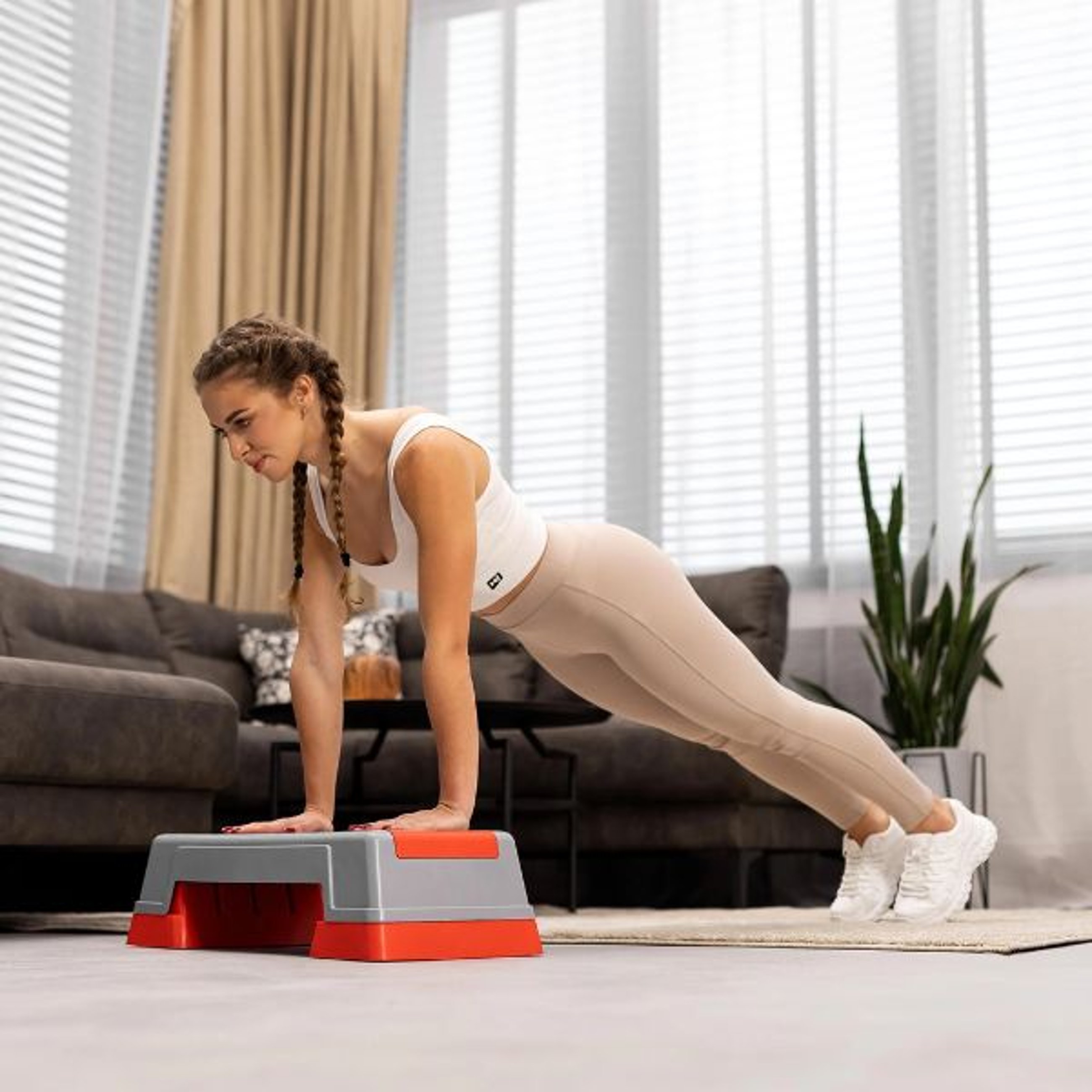 Eine Frau mit geflochtenen Haaren führt eine Plank-Übung auf einem Hop-Sport Aerobic Stepper in Grau und Rot in einem Wohnzimmer aus.