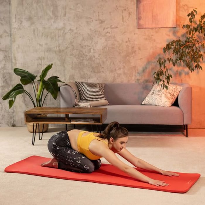 Eine Frau in Sportkleidung führt eine Yoga-Übung auf einer roten Hop-Sport Gymnastikmatte in einem gemütlichen Wohnzimmer durch. Im Hintergrund sind ein graues Sofa, ein Holztisch und Pflanzen zu sehen.