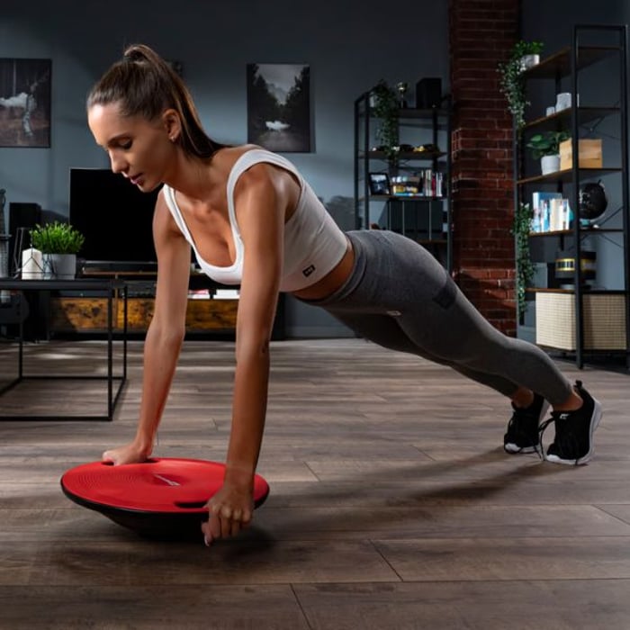 Eine Frau in Sportkleidung führt Übungen mit einem roten Hop-Sport Balance-Board durch. Sie ist in einer Plank-Position und stützt sich mit beiden Händen auf das Board. Im Hintergrund ist ein modern eingerichtetes Wohnzimmer zu sehen.