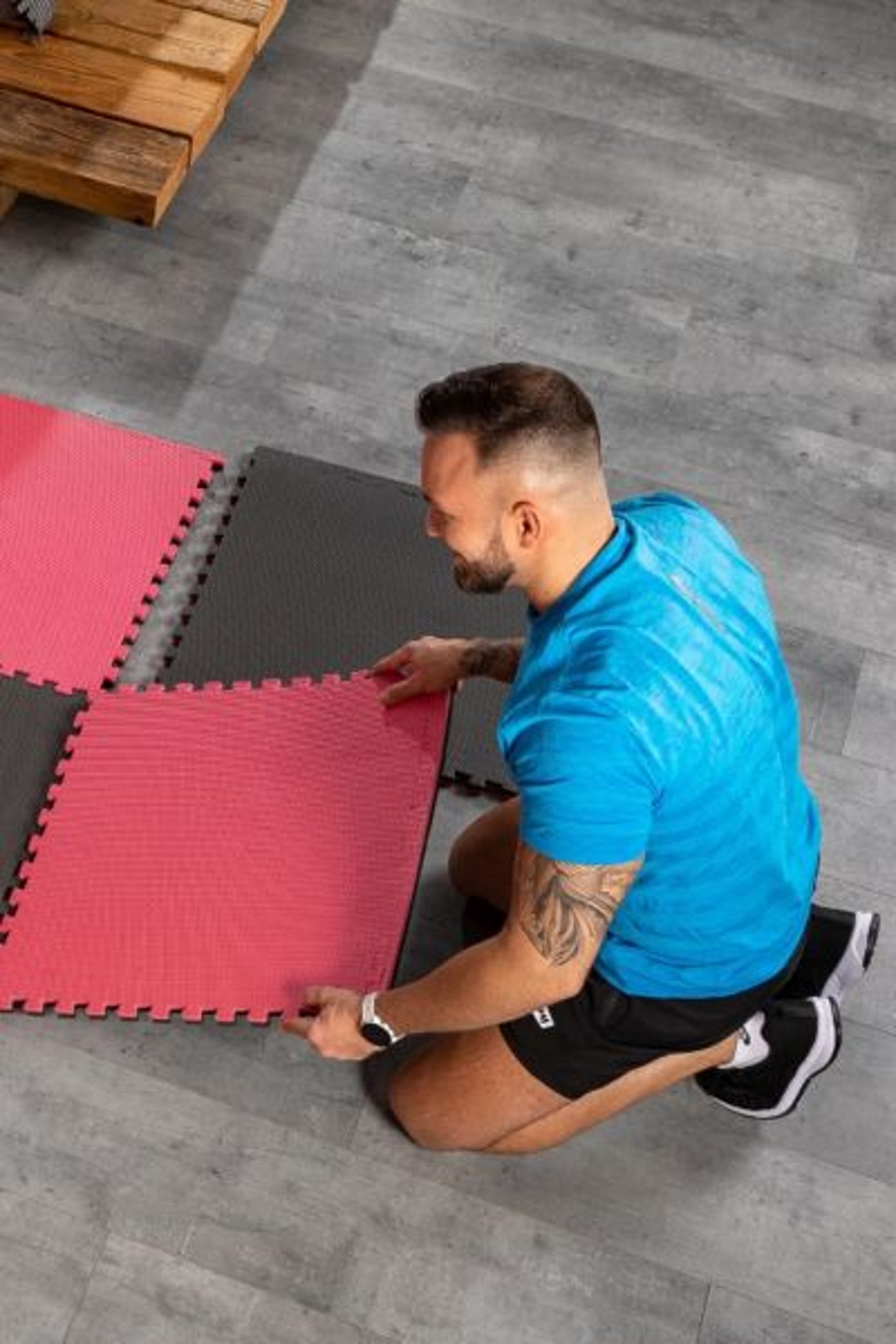 Ein Mann in einem blauen T-Shirt und schwarzen Shorts kniet auf dem Boden und legt eine rote Puzzlematte von Hop-Sport auf eine bereits verlegte schwarz-rote Matte.