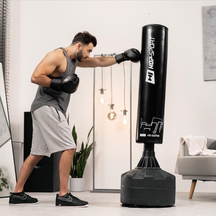 Ein Mann mit Boxhandschuhen schlägt mit seiner rechten Faust auf einen stehenden, schwarzen Hop-Sport Boxsack ein. Er befindet sich in einem modern eingerichteten Raum.